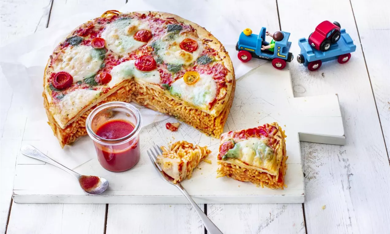 A layered round cake with spaghetti filling, topped with melted cheese, red tomato slices and basil leaves, served on a white board