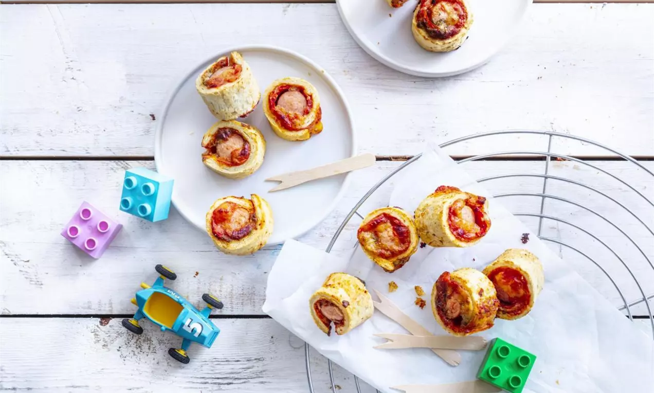 Golden puff pastry rolls filled with sausages, arranged on white plates and parchment paper, surrounded by colorful toy blocks