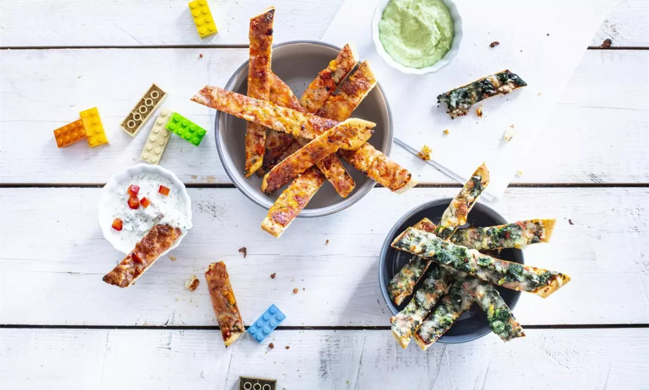 Golden and crisp rectangular pizza sticks with varied toppings placed in bowls on a white wooden surface alongside dipping sauces and colorful toy bricks